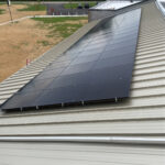 Overhead view of a sloped metal roof with dark solar panels installed, with a parking lot and green landscape in the background.