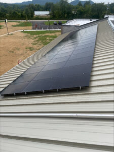 Overhead view of a sloped metal roof with dark solar panels installed, with a parking lot and green landscape in the background.