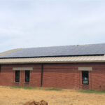 Brick building with a metal roof and solar panels along the eave, a tree on the left, and a clear blue sky above.