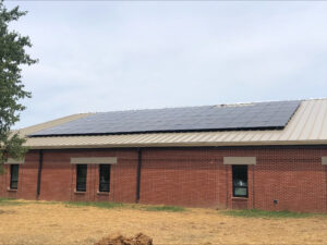 Brick building with a metal roof and solar panels along the eave, a tree on the left, and a clear blue sky above.