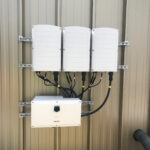 Three white electrical enclosures mounted on a metal frame against a beige wall, with bundled cables leading to a SolarEdge inverter at the bottom.
