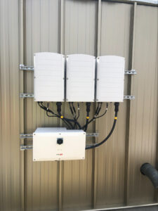 Three white electrical enclosures mounted on a metal frame against a beige wall, with bundled cables leading to a SolarEdge inverter at the bottom.
