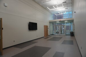 Wide empty hallway in a modern building with a wall-mounted TV on the left and glass exit doors at the far end.