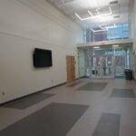 Empty hallway with gray walls, a wall-mounted TV, and glass double doors at the far end beneath an illuminated EXIT sign.