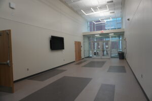 Empty hallway with gray walls, a wall-mounted TV, and glass double doors at the far end beneath an illuminated EXIT sign.