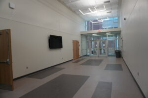 Long modern indoor hallway with gray walls, tiled floor, and large glass doors at the far end; exit sign above the entrance.