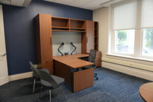 Wood-toned office desk with upper cabinets and storage against a dark blue accent wall, near a large window with blinds.