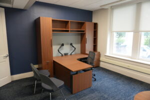Wooden desk with built-in shelving and cabinets against a dark blue wall in a bright office environment.
