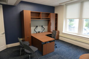 Wooden office desk with built-in storage cabinets and shelves mounted on a blue accent wall, near a large window with blinds.