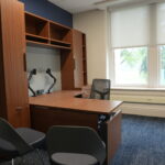Office workspace with dark wood furniture: desks, tall cabinets, and shelving against a blue wall, plus a mesh chair and large window with blinds.