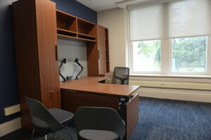Office workspace with dark wood furniture: desks, tall cabinets, and shelving against a blue wall, plus a mesh chair and large window with blinds.