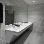 Public restroom sink area: row of three white sinks with chrome faucets on a long counter, large mirror, white subway-tile walls, and a black hand dryer on the left.