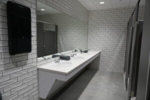 Public restroom sink area: row of three white sinks with chrome faucets on a long counter, large mirror, white subway-tile walls, and a black hand dryer on the left.