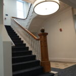 Interior staircase with white balusters, dark-carpeted steps, and a carved wooden newel post under a large circular ceiling light; signs on the wall to the left indicate building directions.