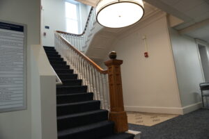 Interior staircase with white balusters, dark-carpeted steps, and a carved wooden newel post under a large circular ceiling light; signs on the wall to the left indicate building directions.