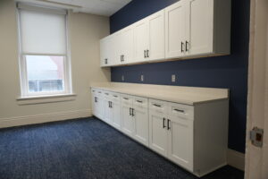 White cabinetry lines the room both above and below a long light countertop, set against a navy wall, with a window and blinds to the left and a blue patterned carpet on the floor; a bright, clean office kitchenette.