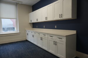 White wall-to-wall cabinets above a long light countertop against a navy blue wall, with a window to the left.