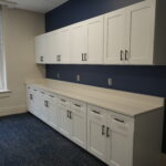 Row of white upper and lower cabinets with black handles along a navy wall in an office kitchenette, window to the left.