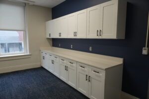 Row of white upper and lower cabinets with black handles along a navy wall in an office kitchenette, window to the left.