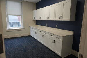 White upper and lower cabinets run along a navy wall with white countertops and a window with a blind on the left side.