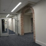 Interior office hallway with glass doors and a brick archway at the end, carpeted floor and overhead fluorescent lighting