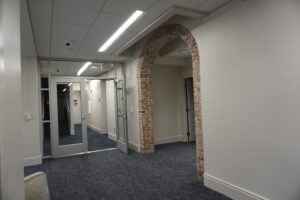 Interior office hallway with glass doors and a brick archway at the end, carpeted floor and overhead fluorescent lighting