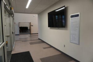 Wide interior hallway with overhead lights, a wall directory sign on the right, and a large wall-mounted screen; an open doorway reveals stacked chairs in a room ahead.