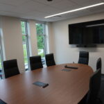 Conference room with an oval wooden table, black chairs, wall-mounted TV and camera, and windows showing trees outside.",