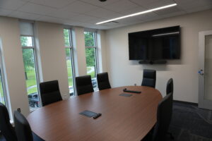 Conference room with an oval wooden table, black chairs, wall-mounted TV and camera, and windows showing trees outside.",