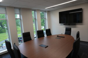 Empty meeting room with a large wood oval table, black chairs, and a wall-mounted flat-screen TV.