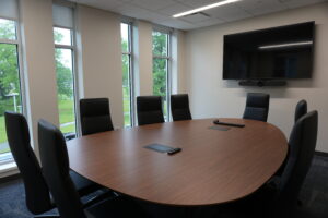 Conference room with a round wooden table, black leather chairs, and a wall-mounted flat-screen monitor on the front wall.