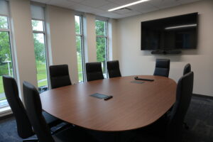 Conference room with a large round wooden table, black leather chairs, and a wall-mounted flat-screen TV.