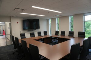 Modern conference room with a large U-shaped wooden table, black chairs, and a wall-mounted TV with a video camera below it; windows show greenery outside.