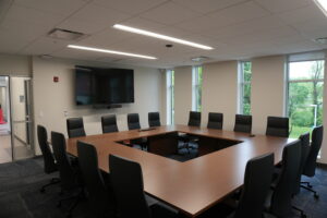 Large conference room with U-shaped wooden tables and numerous black chairs, a wall-mounted flat-screen display with a video conference camera beneath it, and tall windows showing greenery outside.