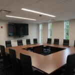 Conference room with a large wooden U-shaped table, black leather chairs, a wall-mounted TV, and video conferencing camera below it in front of windows showing greenery.