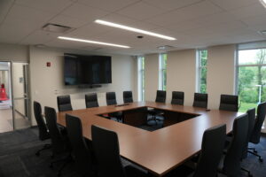 Conference room with a large wooden U-shaped table, black leather chairs, a wall-mounted TV, and video conferencing camera below it in front of windows showing greenery.