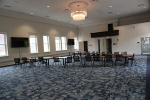 Large conference room with long U-shaped tables and black chairs, windows along the walls, and a central chandelier overhead