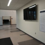 Hallway in a modern building with a wall-mounted flat-screen monitor and a vertical directory sign labeled Administration Building on the right; doorway with stacked chairs visible ahead.