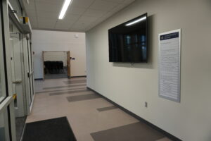 Hallway in a modern building with a wall-mounted flat-screen monitor and a vertical directory sign labeled Administration Building on the right; doorway with stacked chairs visible ahead.