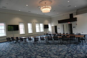 Spacious meeting room with long rectangular tables arranged in a U shape, black chairs, and a large chandelier overhead.