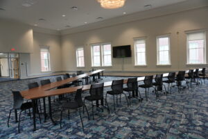 Empty conference room with long wooden tables arranged in a U shape, black mesh chairs, and a wall-mounted TV against the far wall.] ,