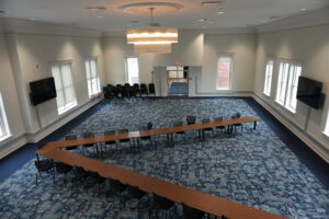 Spacious conference room with a U-shaped wooden table setup, black chairs, and a patterned blue carpet under a large central chandelier.
