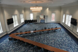 A large conference room with U-shaped wooden tables and black chairs, chandeliers overhead, and tall windows along the walls (empty room).