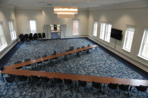 Bright, empty conference room with long, U-shaped wooden tables and black chairs; additional stacked chairs along the wall and a large chandelier overhead.