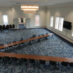 Empty conference room set up in a U-shaped table arrangement with black chairs and a blue patterned carpet, large circular chandelier overhead.