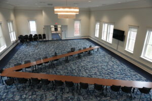 Empty conference room set up in a U-shaped table arrangement with black chairs and a blue patterned carpet, large circular chandelier overhead.
