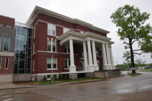 Brick building with a white-columned portico and modern glass extension on the left, under a cloudy sky.