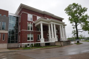 Red brick building with a white-columned entrance and a glass-sided extension on the left, beside a large green tree.
