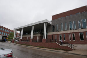 Campus building exterior with a covered entrance, white columns, brick and gray panel facade, under an overcast sky.