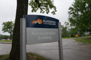Sign for Bluegrass Community & Technical College with orange sun logo; Administration Building visible, trees and road in the background.